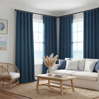 A set of deep navy blue grommet curtains hanging on a black rod in a bright, modern living room with a white sofa, wooden coffee table, and ocean view windows.