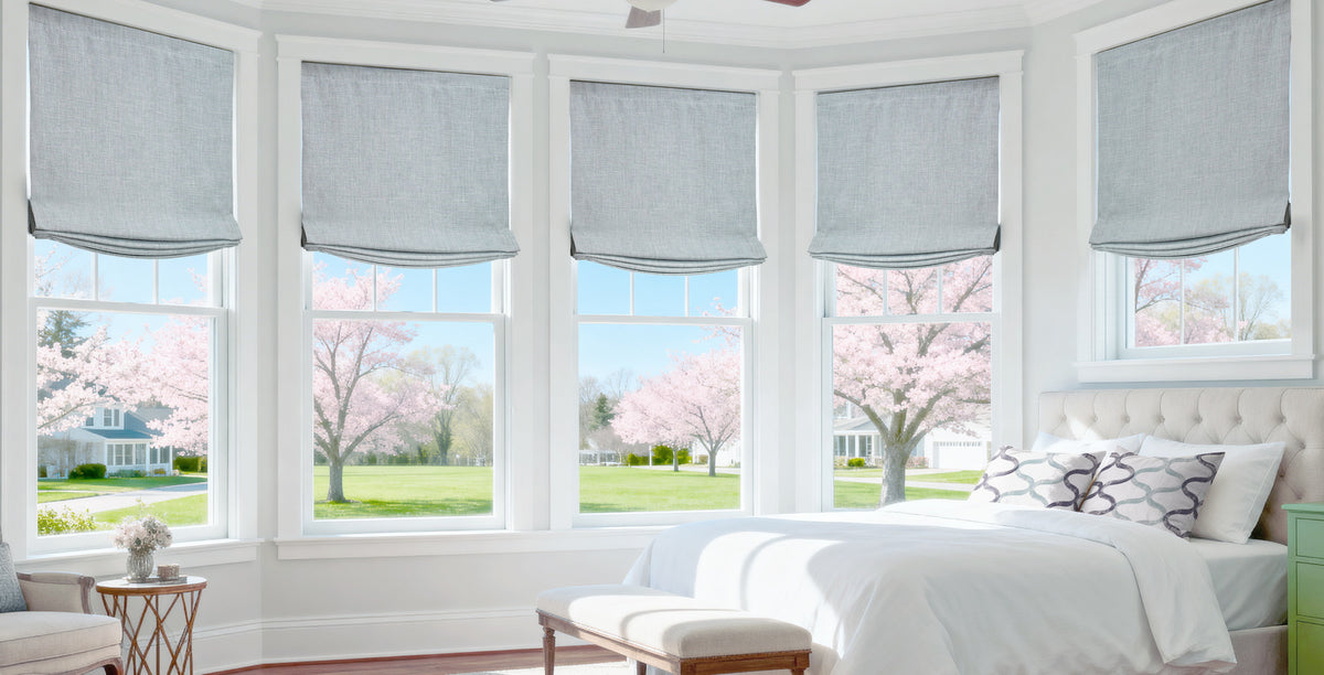 Light gray custom Roman shades dress a bright bay window bedroom, offering elegant light filtering, soft texture, and a clean tailored look with views of blooming spring trees.