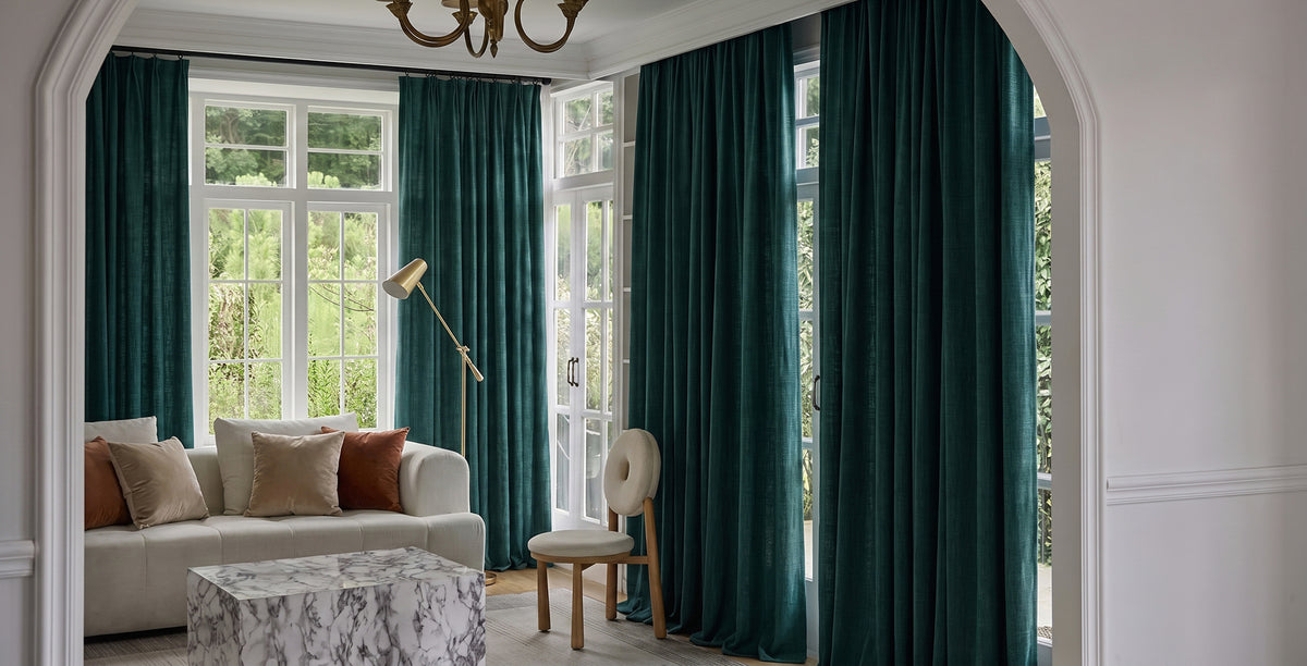 Mauve velvet curtains frame tall arched windows in an elegant bedroom with soft neutral furnishings and blush accents.