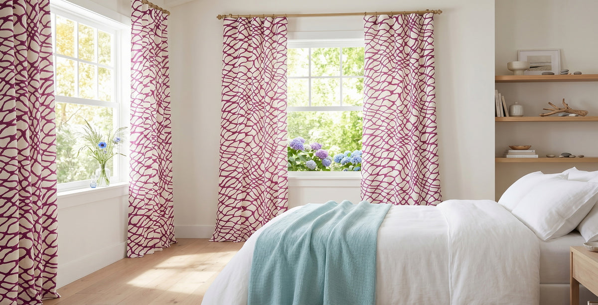 Bright, airy bedroom featuring white bedding and light wood floors, styled with bold pink abstract-patterned cotton curtains on three sunlit windows. Natural light fills the space, highlighting soft neutral walls, floating wood shelves with minimalist décor, and a pale blue throw blanket draped across the bed. Green foliage and blooming hydrangeas are visible outside the windows, creating a fresh, inviting atmosphere.