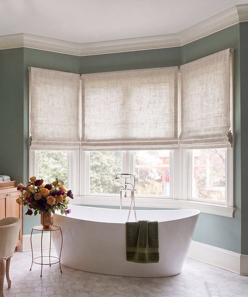 Gravity Italian Faux Linen: Soft ivory Roman shades filter natural light across four tall bathroom windows surrounding a freestanding tub.