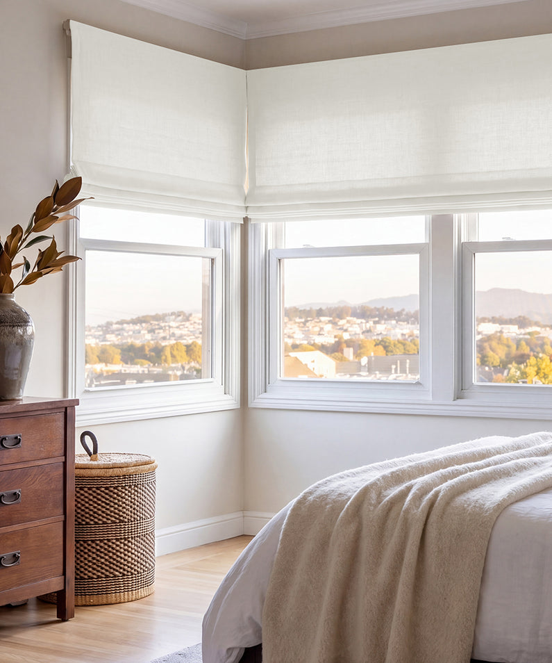 Magnolia Off White: Off-white faux linen Roman shades softly filter sunlight above a neutral sofa accented with warm, earthy pillows.