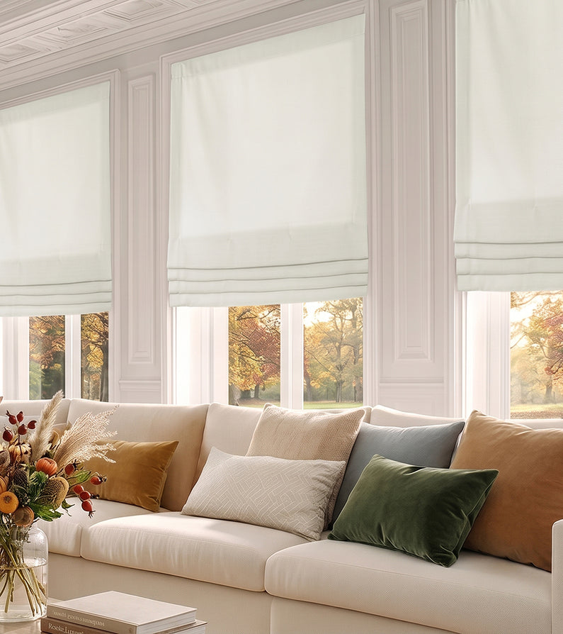 Magnolia Off White: Off-white faux linen Roman shades softly filter sunlight above a neutral sofa accented with warm, earthy pillows.