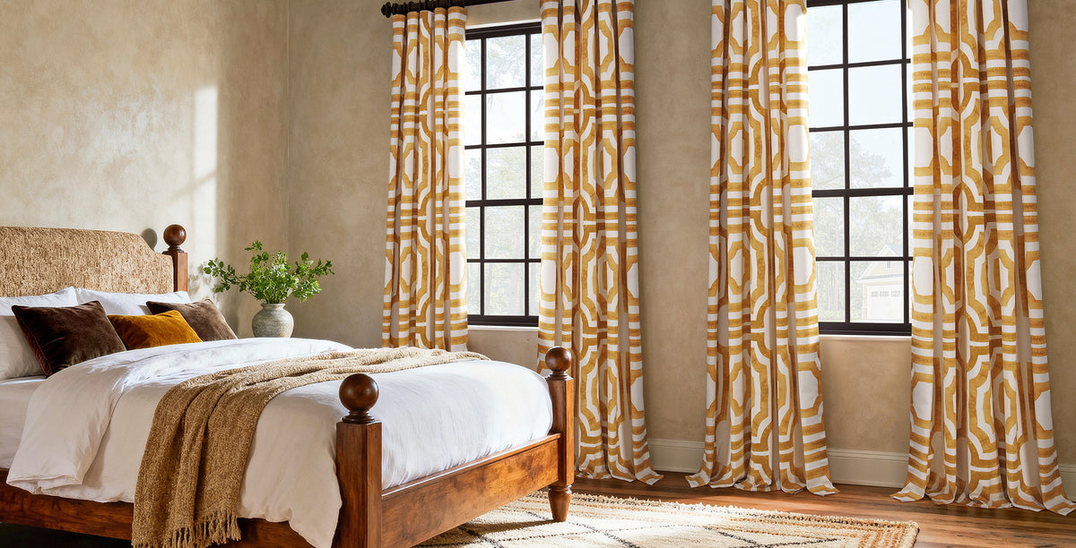 Mecca Gold patterned curtains framing tall bedroom windows with a wood bed and warm neutral décor.
