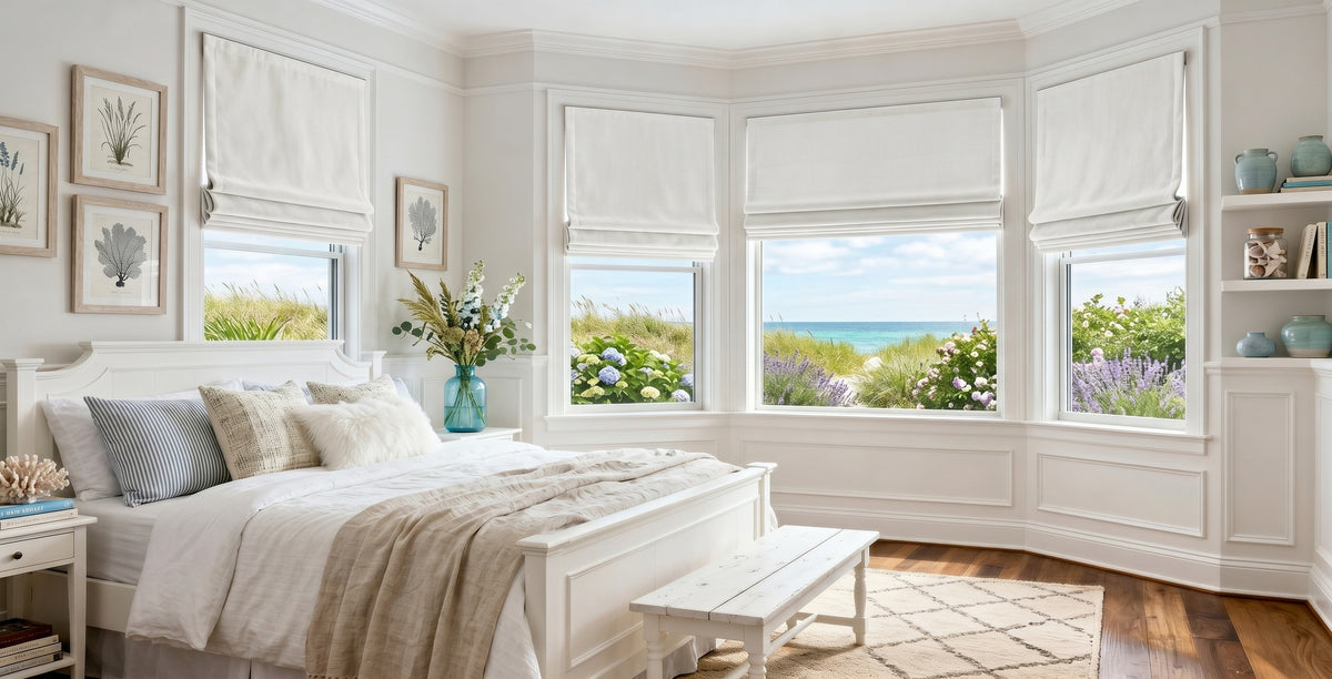 Coastal-inspired bedroom featuring custom white Roman shades on a bay window overlooking beach dunes and ocean views. The bright, airy space includes white paneled walls, crisp white bedding layered with neutral textiles, botanical wall art, and soft natural light. Fresh flowers and coastal décor accents create a serene, relaxed retreat.