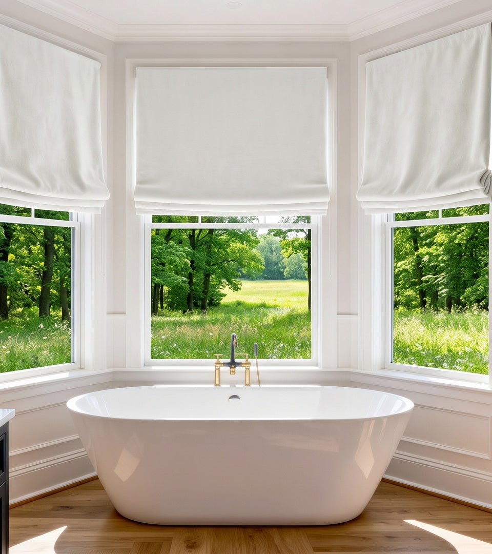 White Roman shades covering bathroom bay windows above a freestanding tub with peaceful garden views.