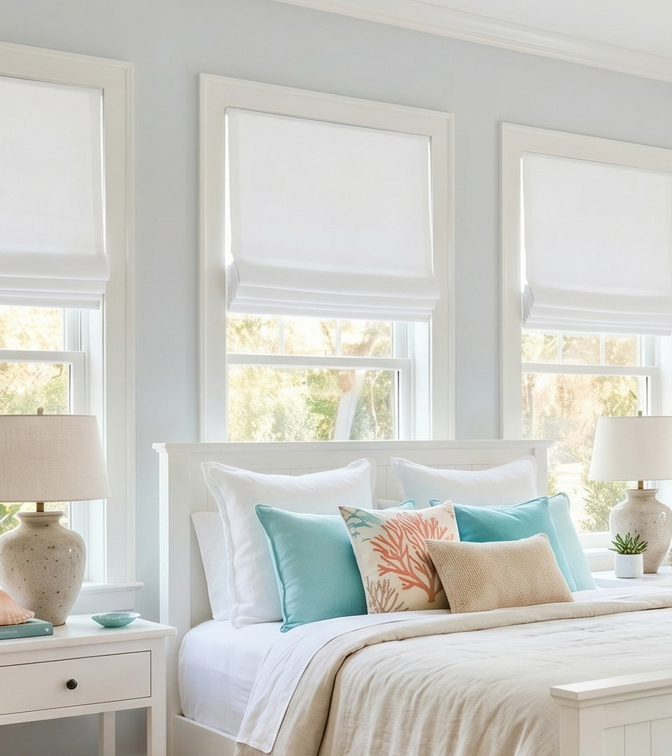 White custom Roman shades installed in a bright bedroom with large windows, layered bedding, decorative accent pillows, and soft natural light filtering into the space.